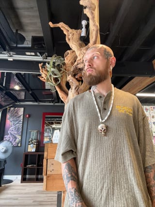 Ryan wears a grey shirt and skull pendant beside a driftwood display in the studio.