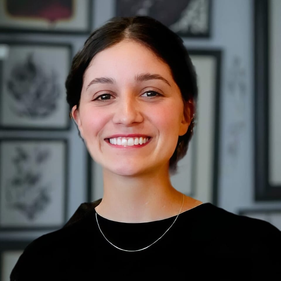 Stellie smiles in a black top and necklace in front of framed artwork.