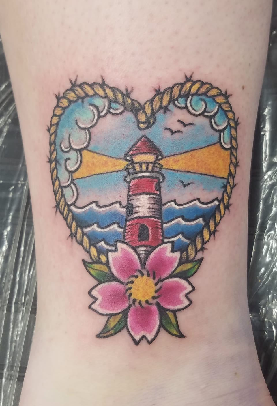 A lighthouse and pink flower sit inside a heart-shaped seaside tattoo.