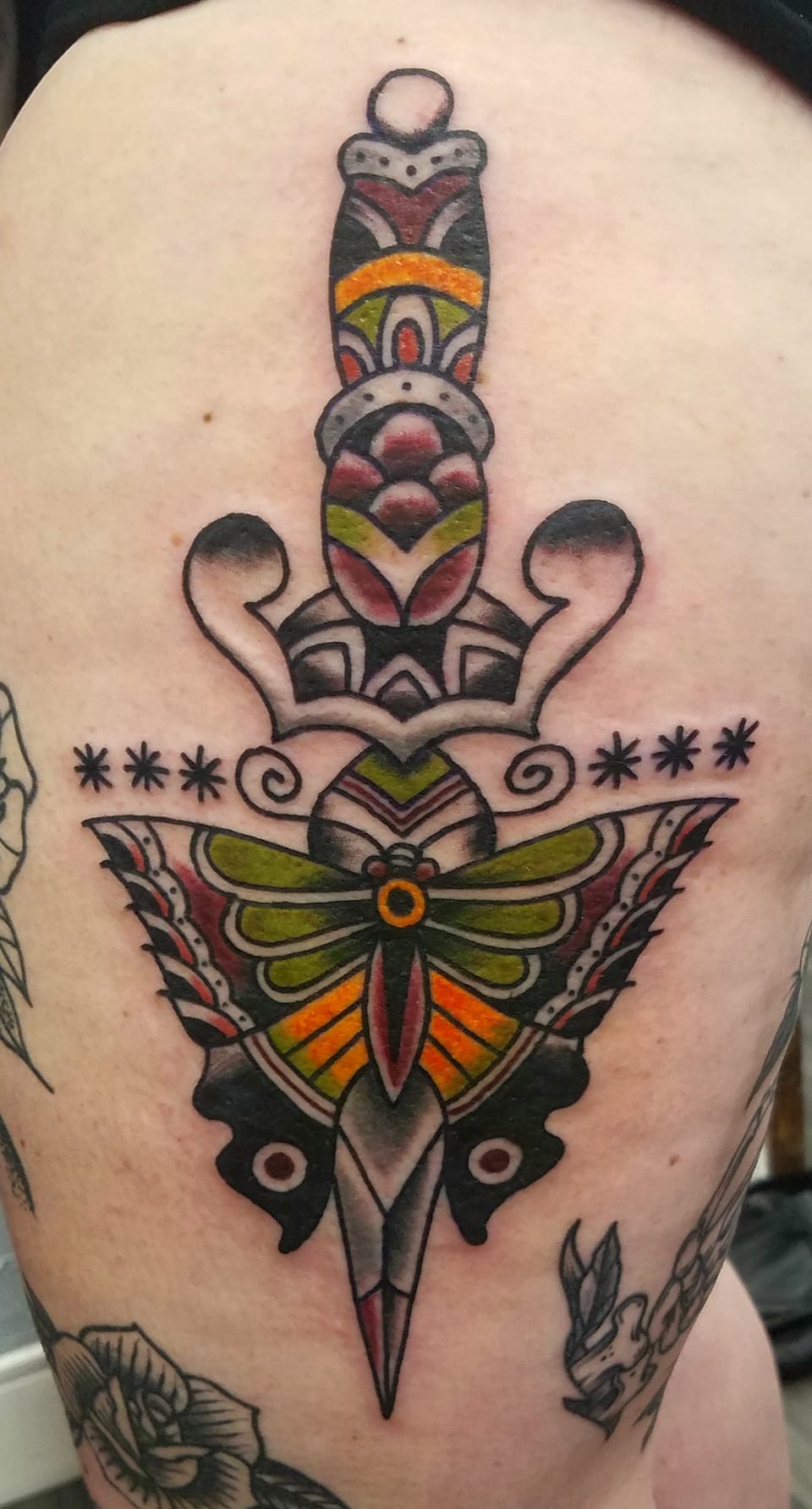 A colorful butterfly spreads its wings across a decorated dagger tattoo.