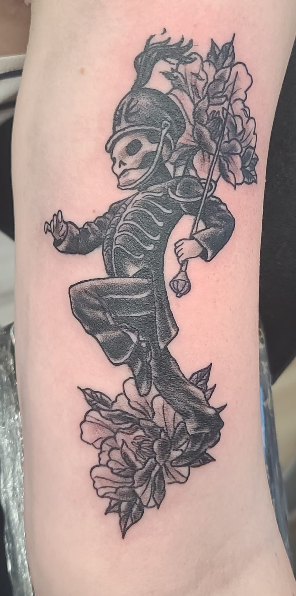 A skeleton in a feathered helmet steps between flowers in a black tattoo.