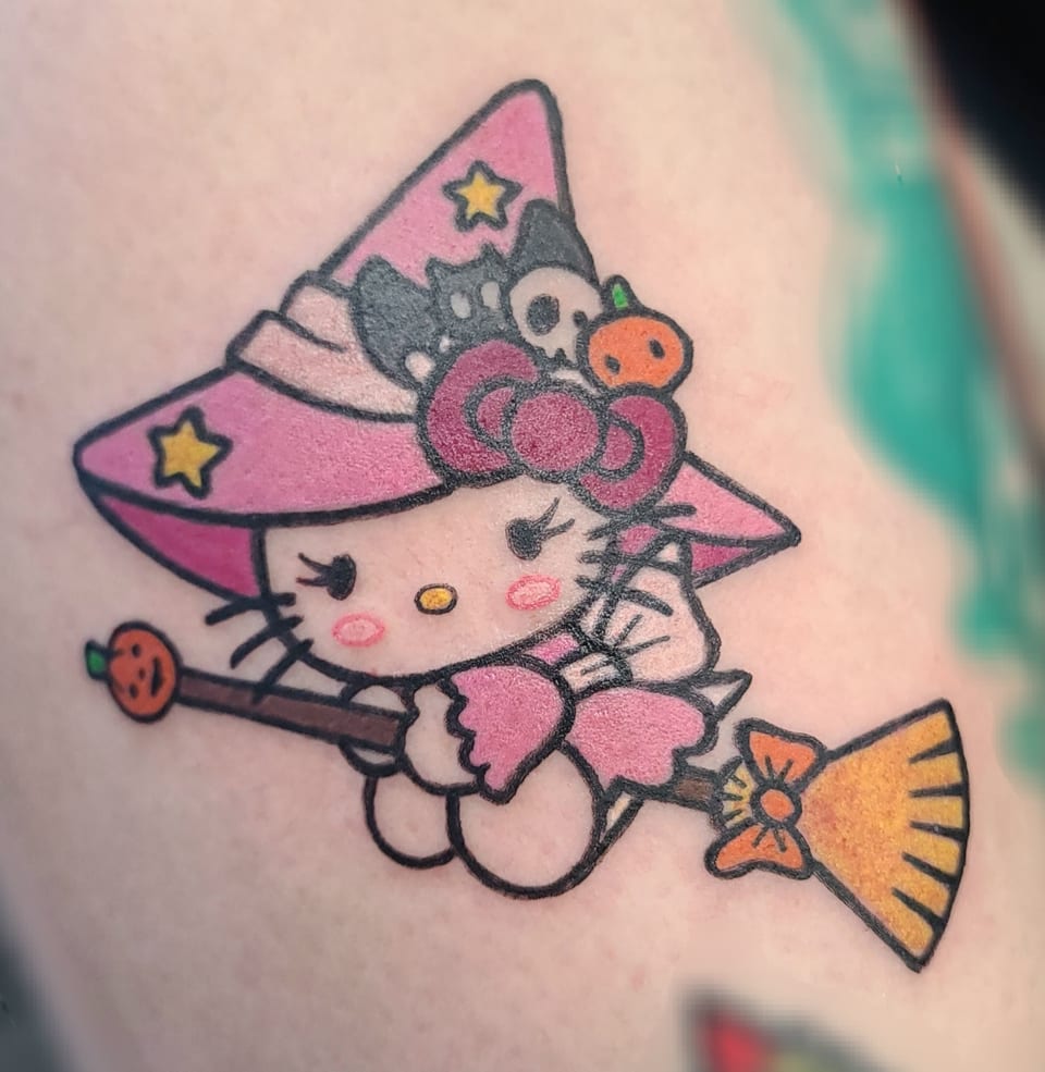 A pink-hatted cartoon cat rides a broom in a colorful tattoo.