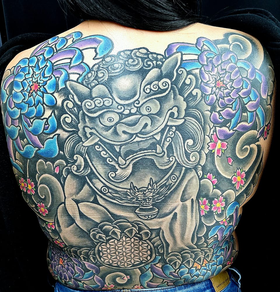A large blue guardian-lion tattoo with flowers covers the upper back.