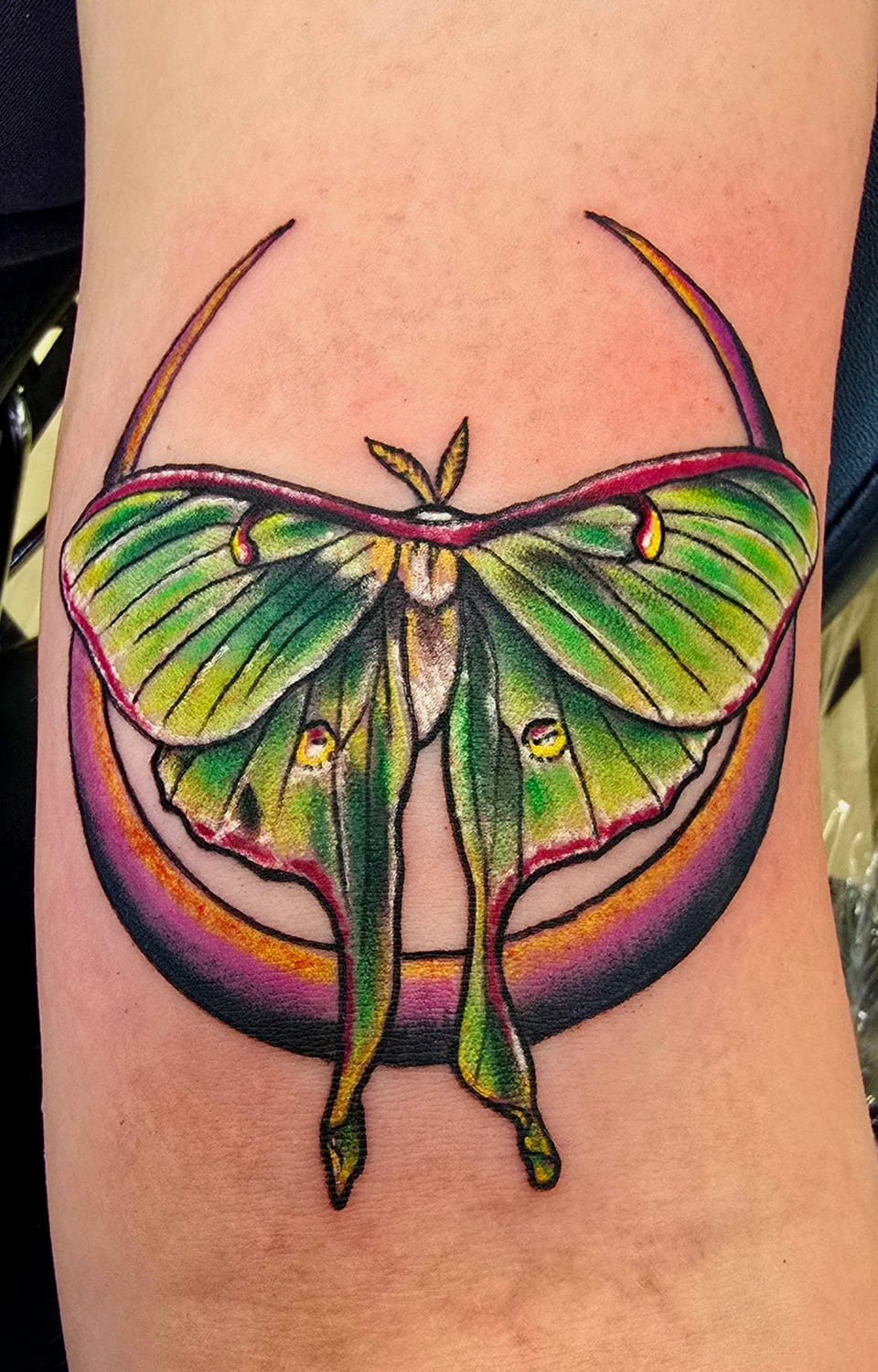 A green moth rests against a purple crescent in a colorful tattoo.