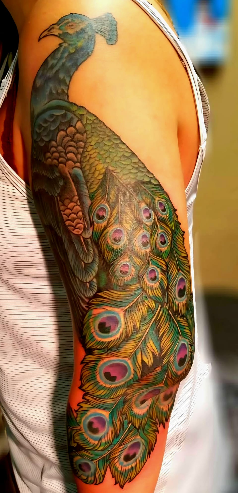 A blue and green peacock tattoo trails patterned feathers down an arm.