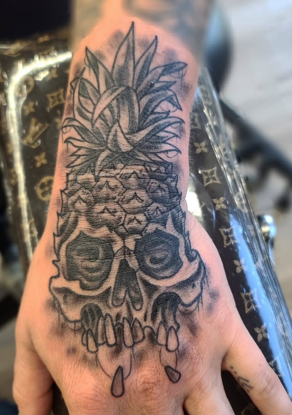 A skull topped with a pineapple forms a black and grey hand tattoo.