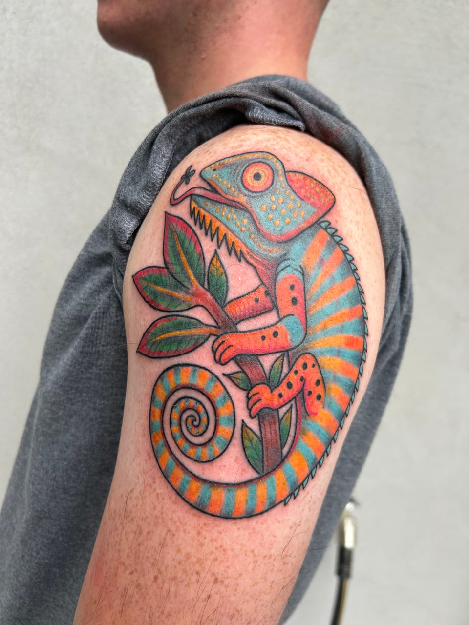 An orange and blue chameleon tattoo curls around green leaves on the upper arm.