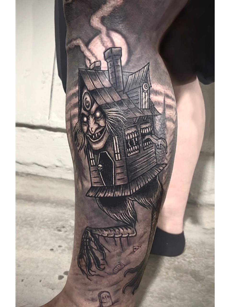 A black and grey haunted-house tattoo has a face, a steep roof and a long clawed leg.