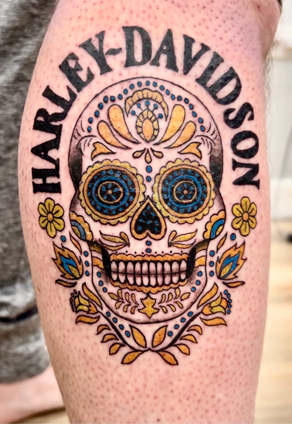 A yellow and blue sugar-skull tattoo sits beneath curved black lettering.