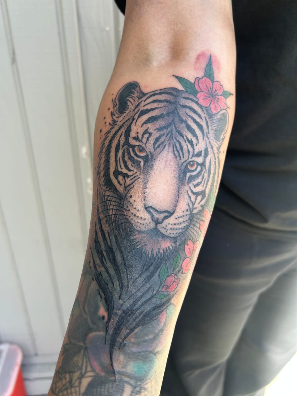 A shaded tiger face tattoo follows the forearm beside a pink flower.