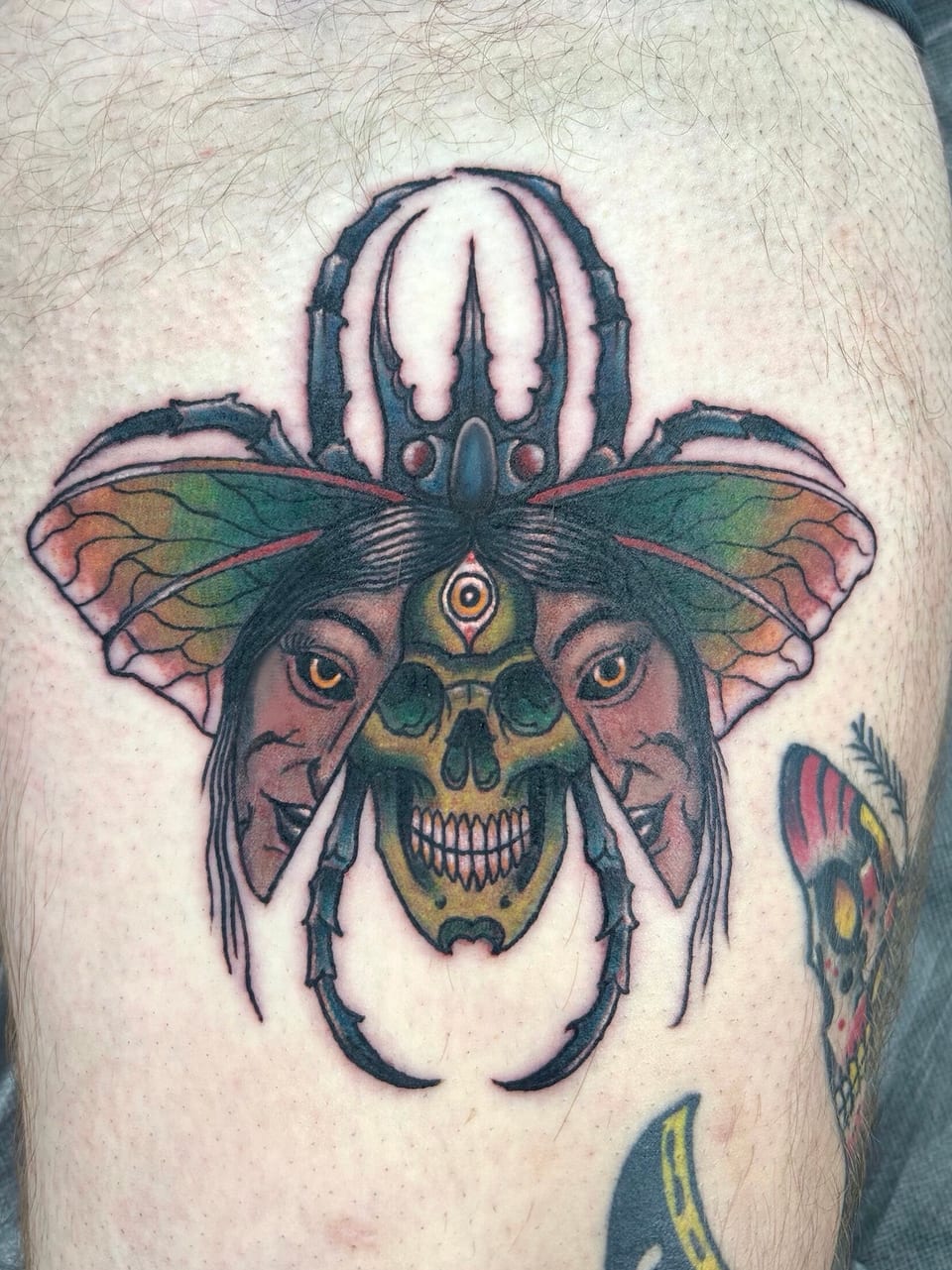 A winged beetle tattoo combines a skull with eyes and colored wing patterns.