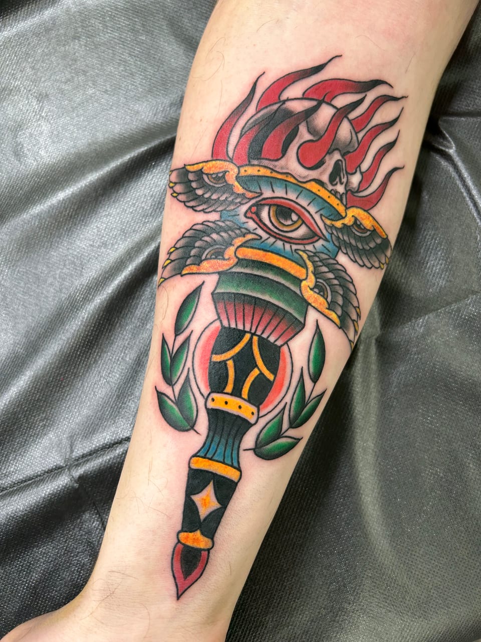A colorful dagger tattoo passes through an eye beneath wings and a flaming skull.