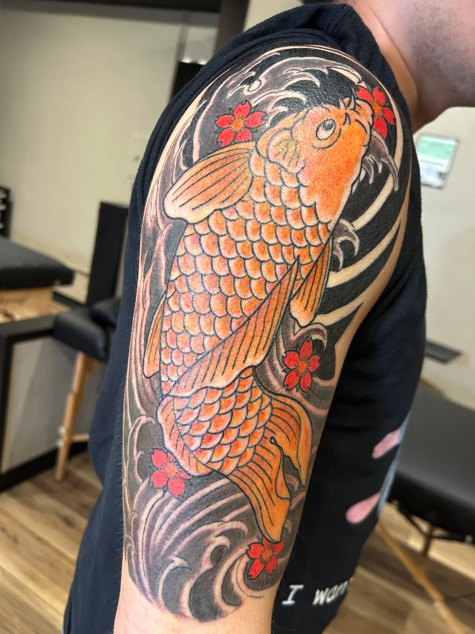 An orange koi tattoo swims through black waves and red flowers on the upper arm.