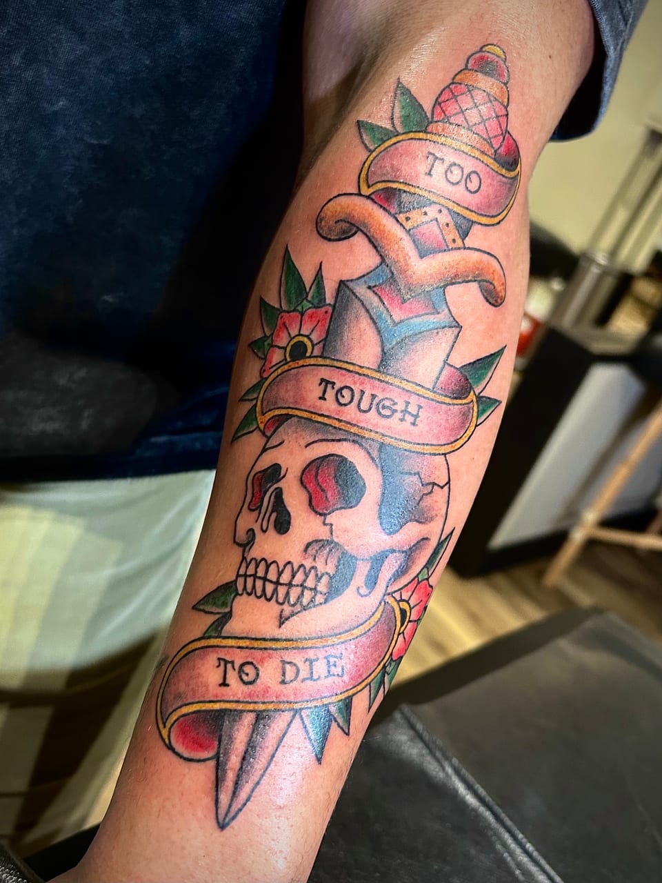 A dagger, skull, flowers and banners form a colorful forearm tattoo.
