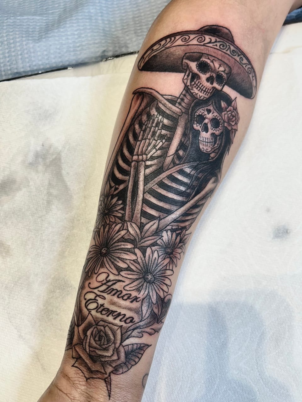 A black and grey skeleton couple tattoo sits above flowers and a lettering banner.