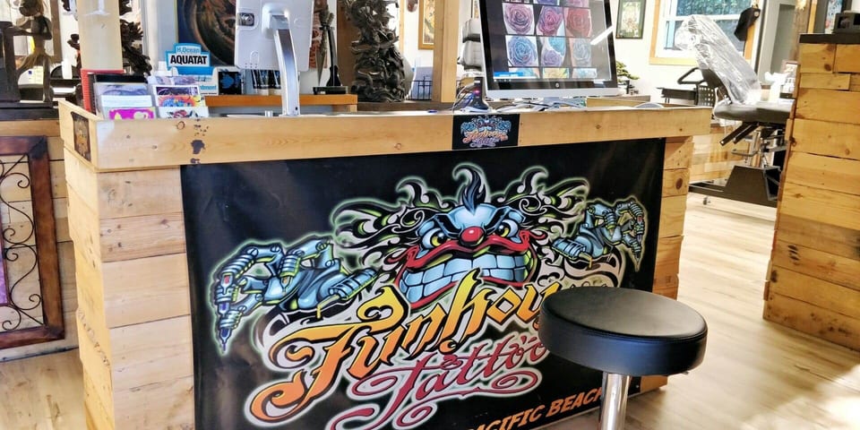 A wooden reception counter has a Funhouse Tattoo clown banner across its front and monitors on top.