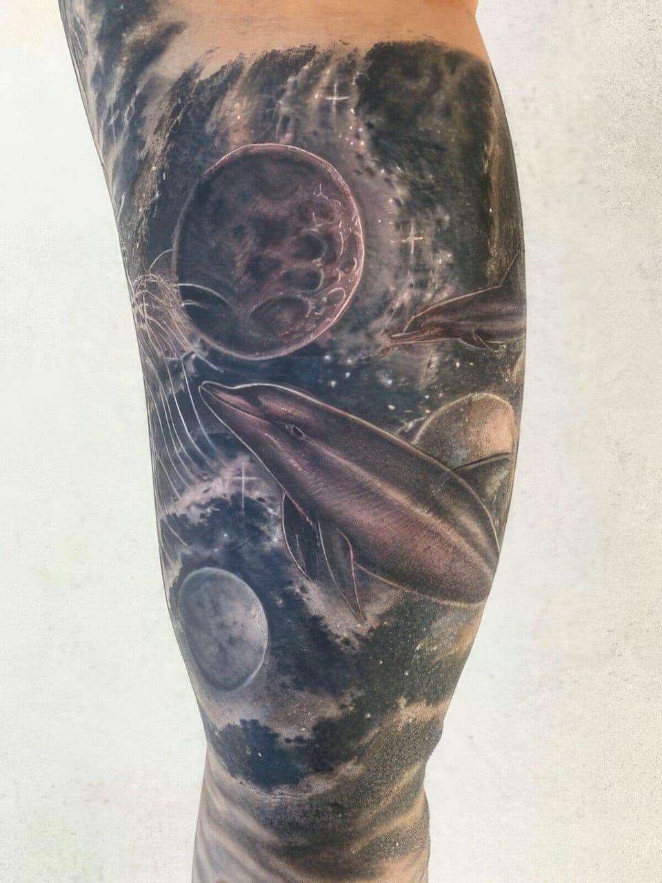 A black-and-grey realistic dolphin and planets tattoo covers an arm.