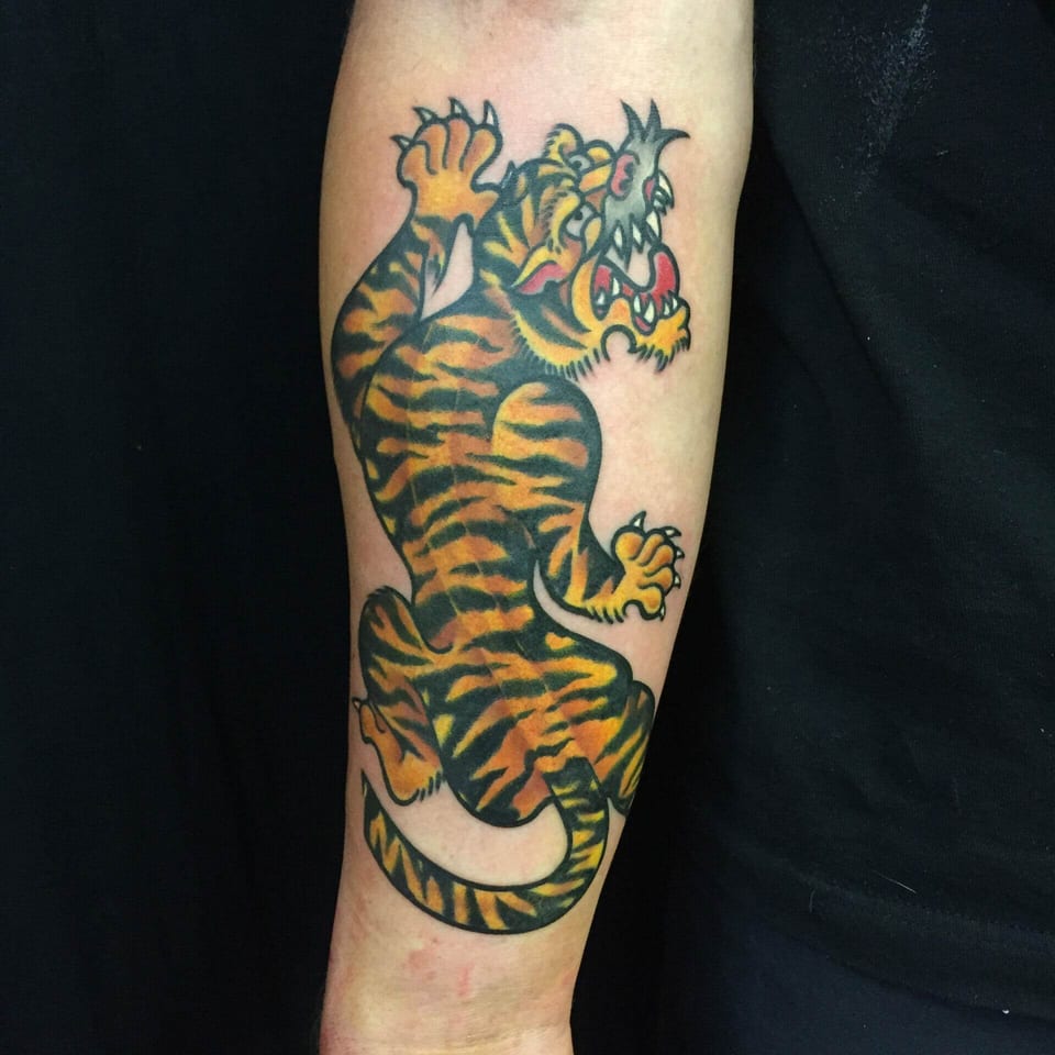 A traditional orange tiger with black stripes and raised paws stretches down a forearm.