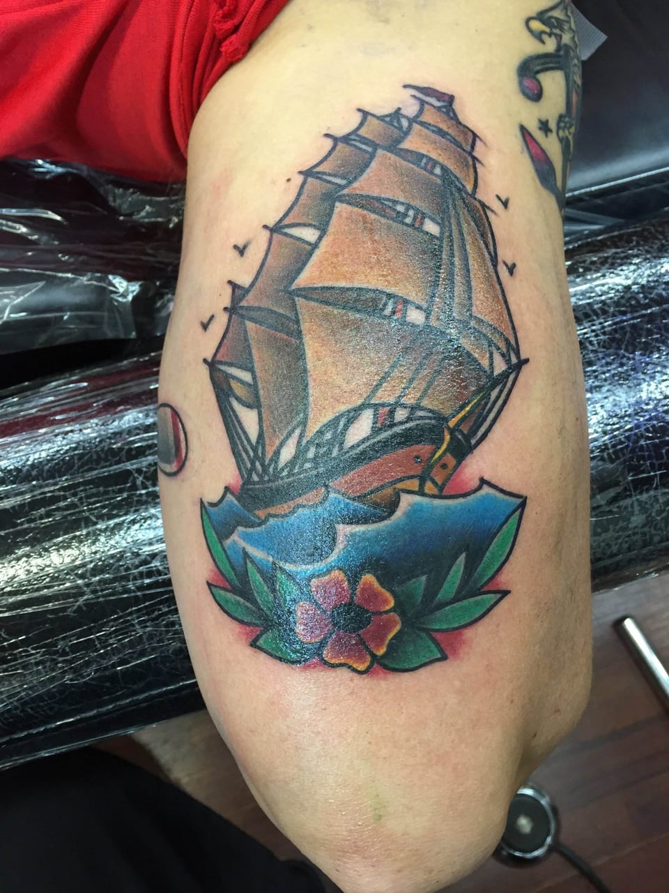 A traditional sailing ship on blue waves with a red flower covers an upper arm.