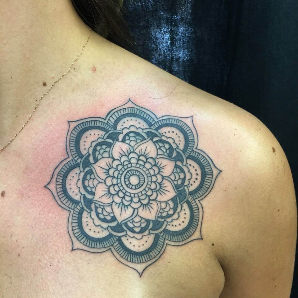 A black geometric mandala with layered petals and dotwork sits below a collarbone.