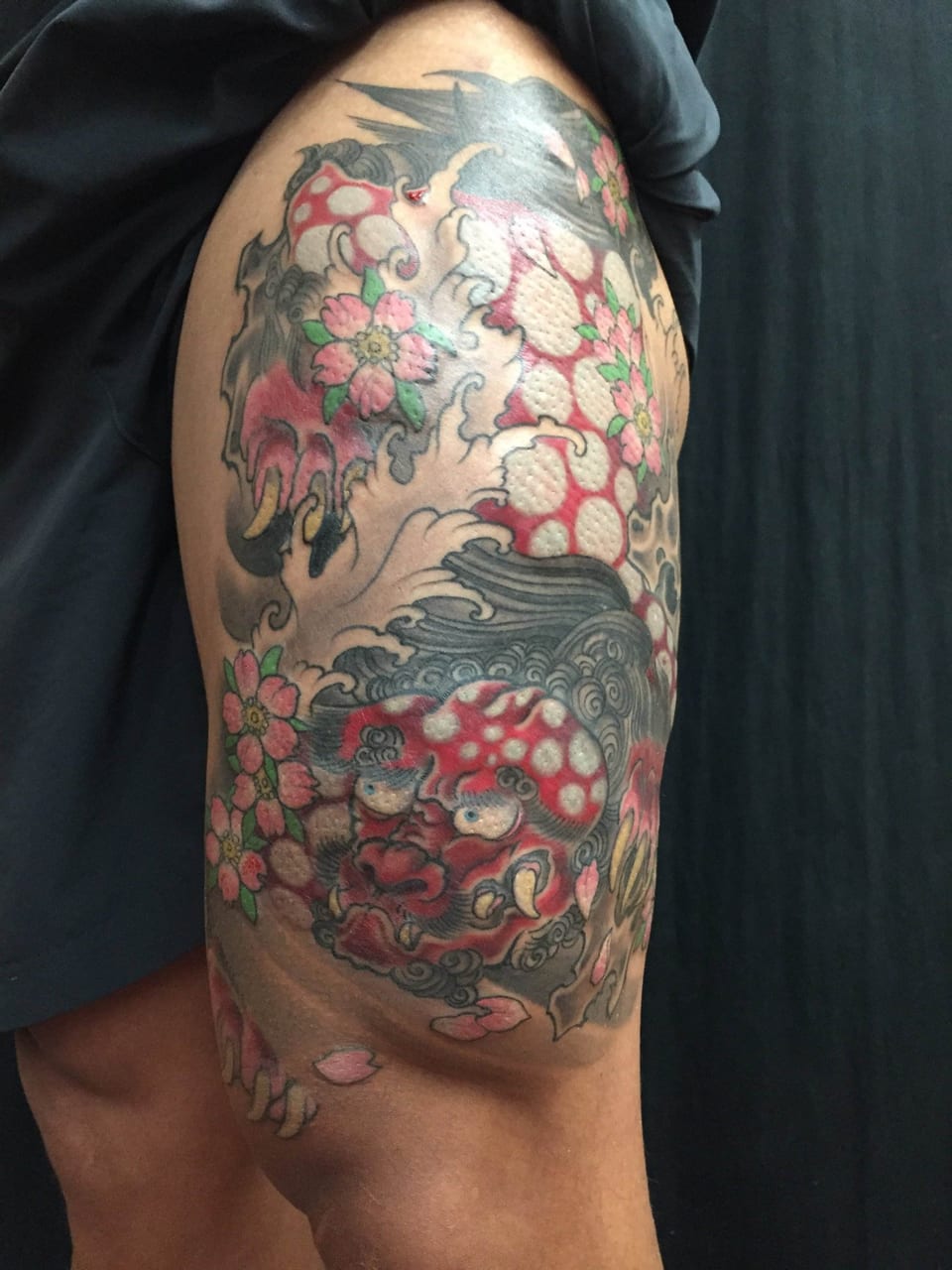 A Japanese red foo dog with pale spots, pink blossoms and grey waves covers a thigh.