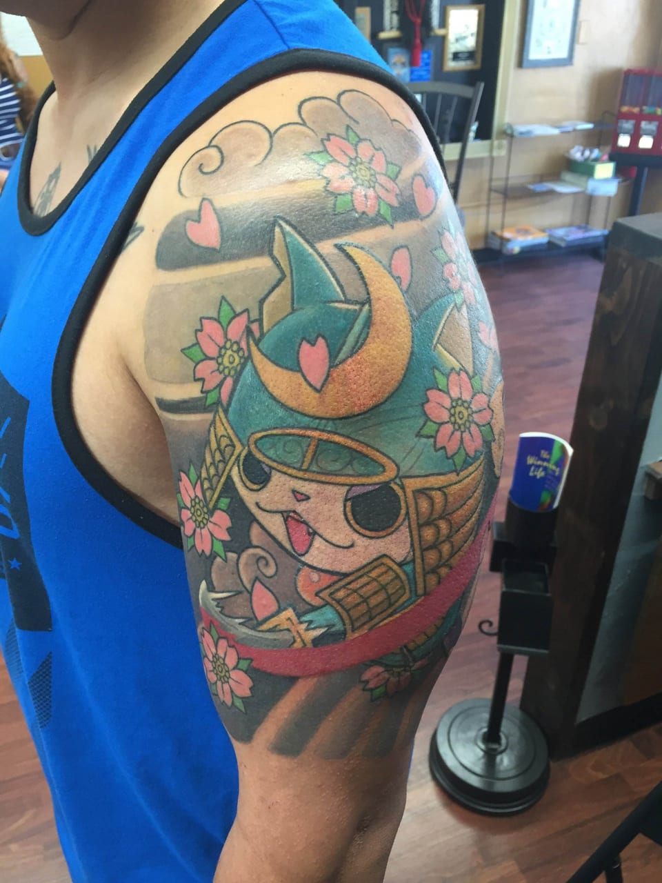 A colorful Japanese-style samurai cat with a teal helmet, hearts and blossoms covers an upper arm.