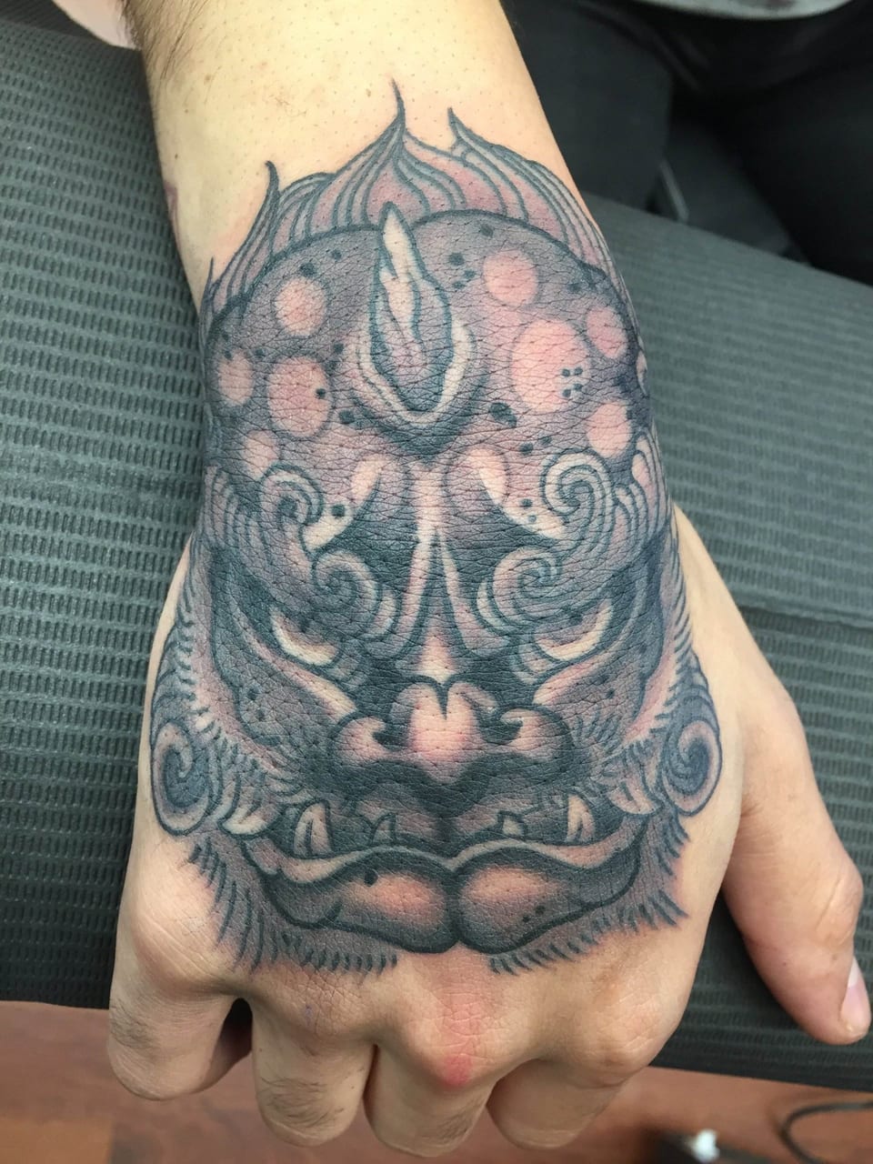 A black-and-grey Japanese foo dog face with curling details covers the back of a hand.