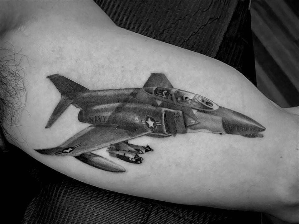 A black-and-grey realistic fighter jet stretches across an inner upper arm.