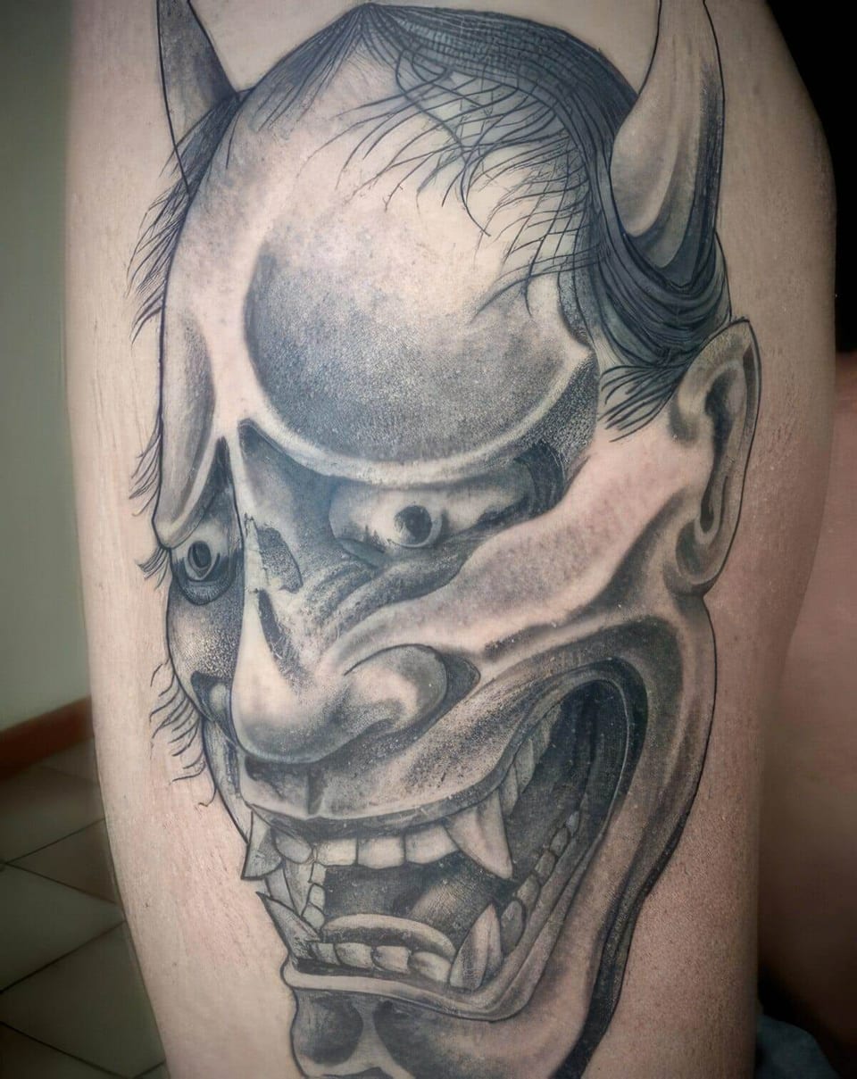 A black-and-grey Japanese demon mask with long horns and sharp teeth covers a thigh.