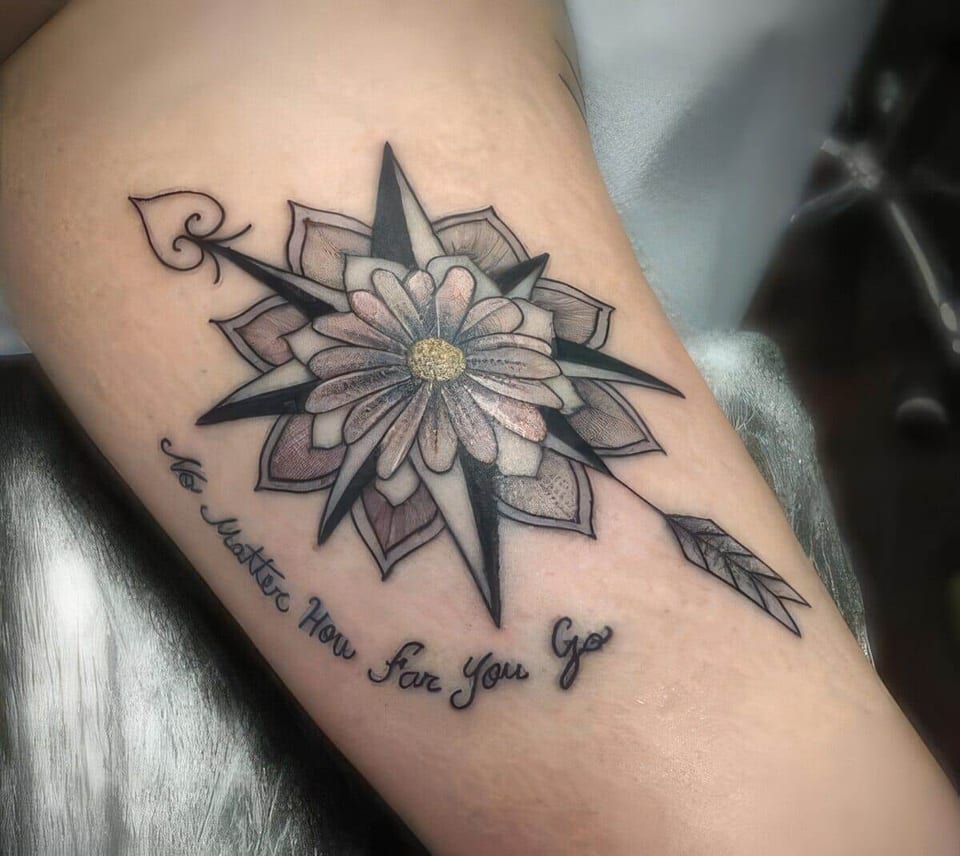 A black-and-grey illustrative compass flower, arrow and curved script cover a forearm.