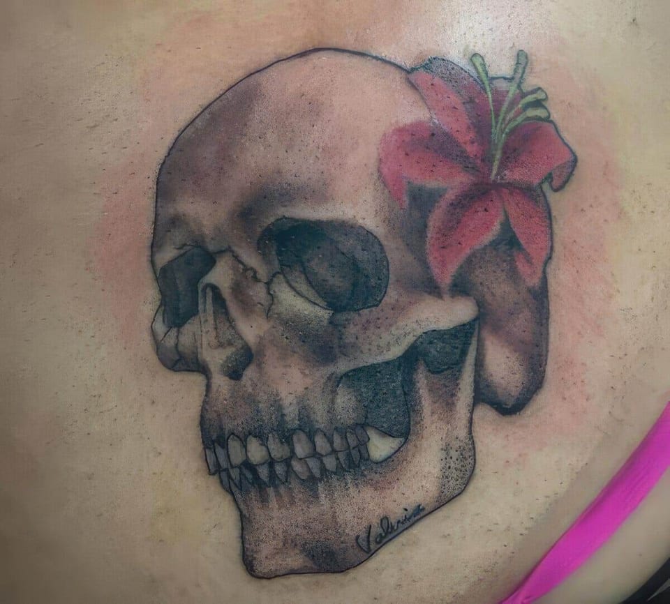 A black and grey realistic skull with a pink lily tattoo covers the shoulder blade.