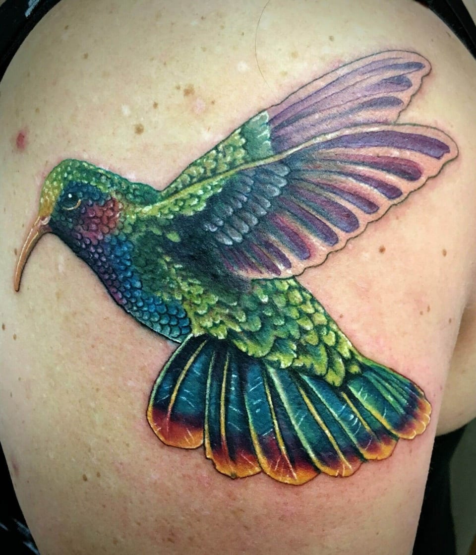 A realistic green, blue and purple hummingbird with spread wings covers a shoulder.