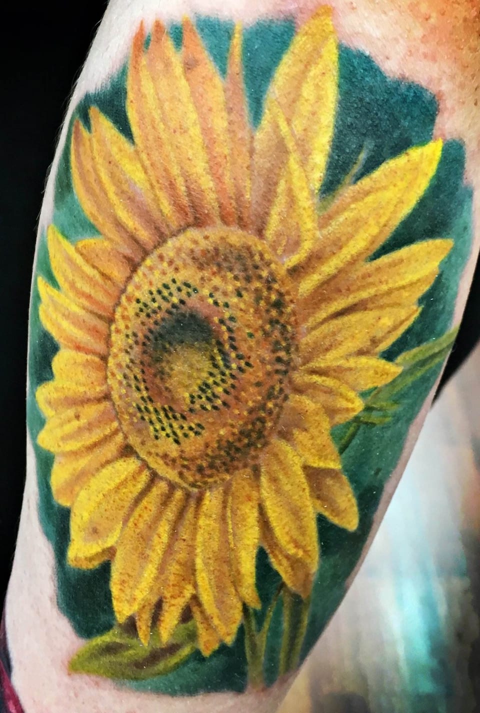A realistic yellow sunflower tattoo with a dark green background covers an arm.