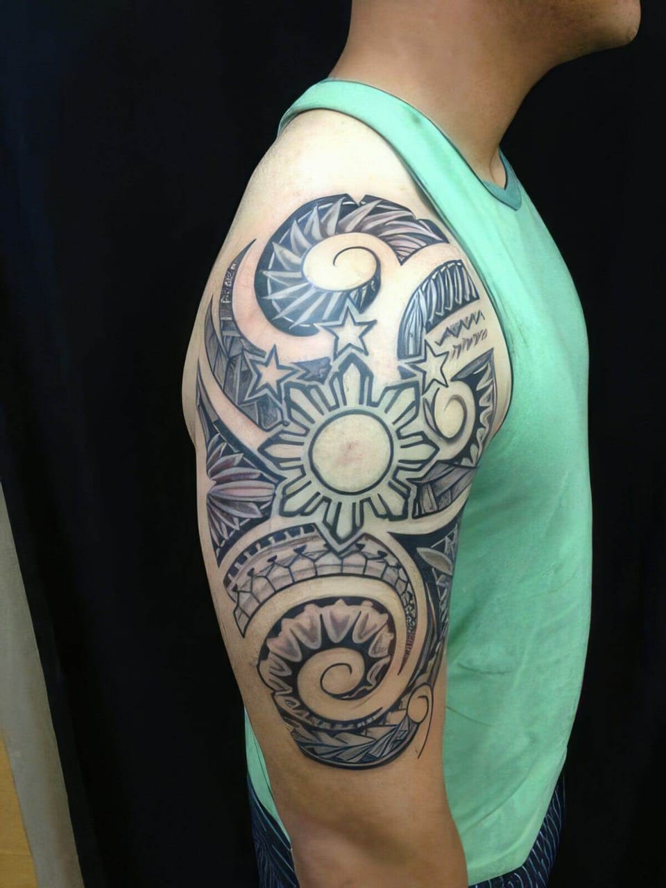 Black and grey Polynesian-style tribal tattoo with a sun emblem on the upper arm of a man in a green tank top, tattooed by Seth Reynolds