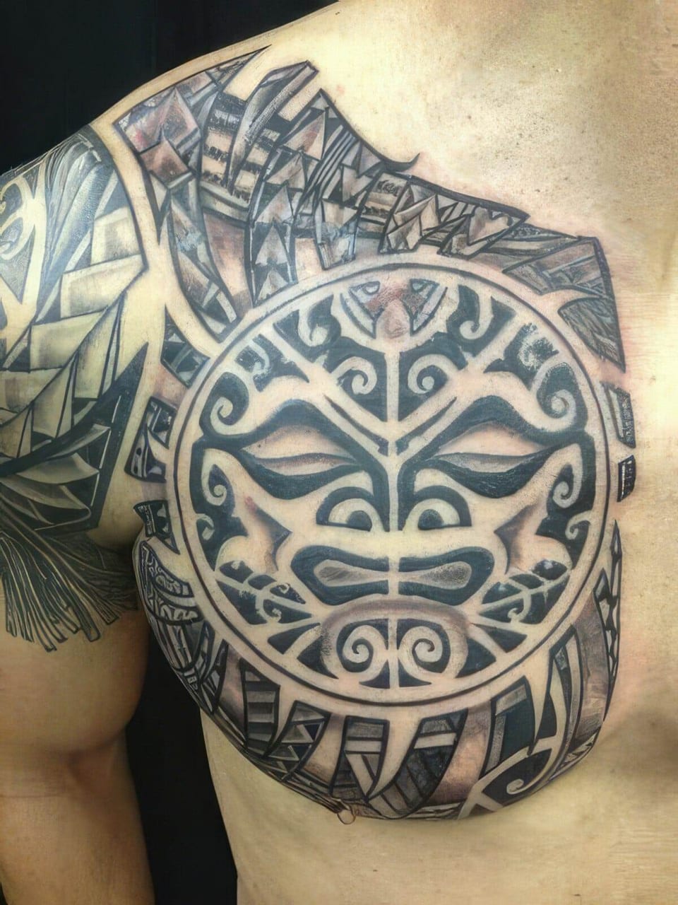 Black Polynesian-style sun mask with surrounding patterns tattooed on a chest and shoulder, tattooed by Seth Reynolds