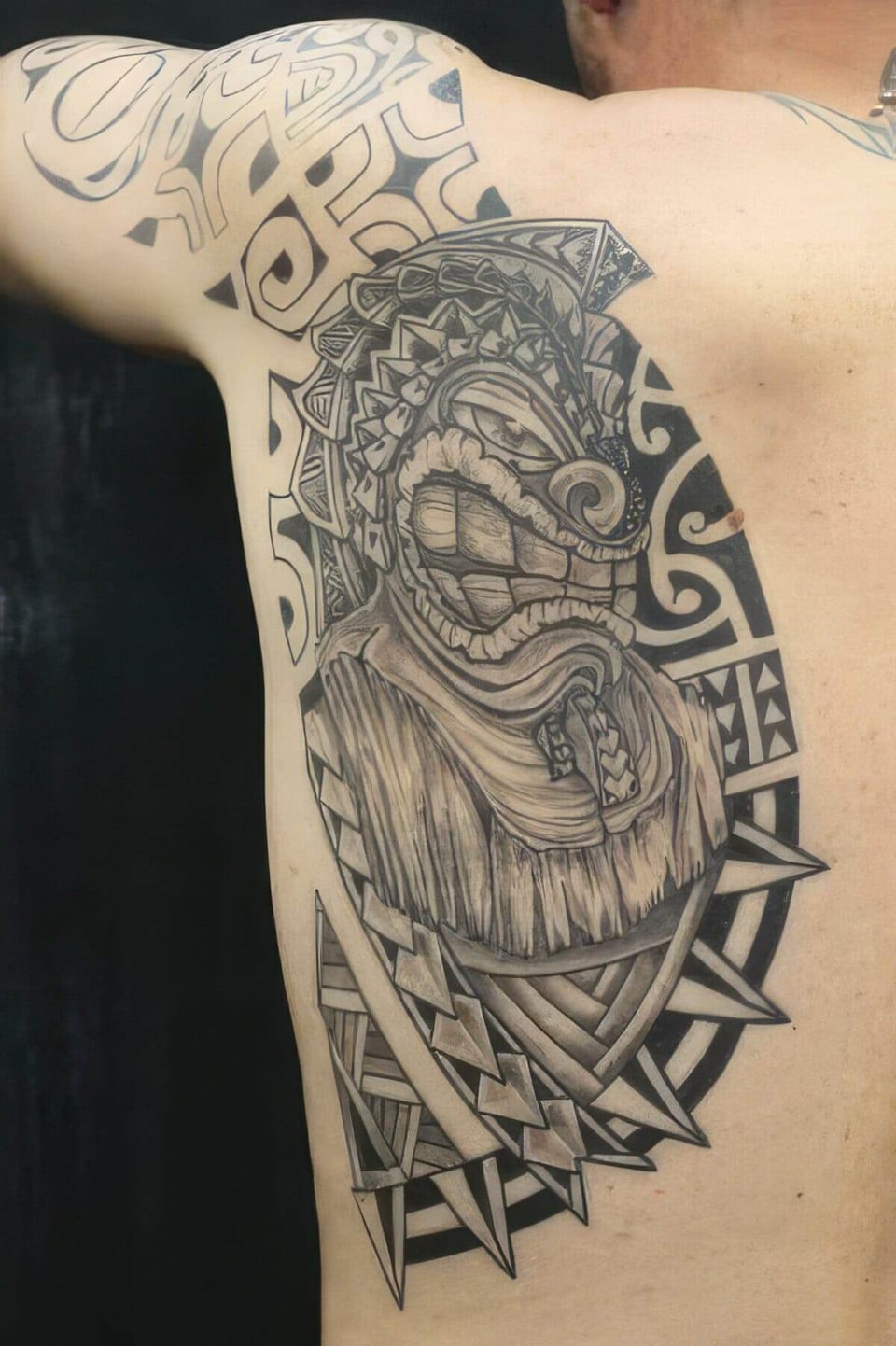 Black and grey Polynesian-style mask and geometric patterns tattooed on a shoulder and chest, tattooed by Seth Reynolds