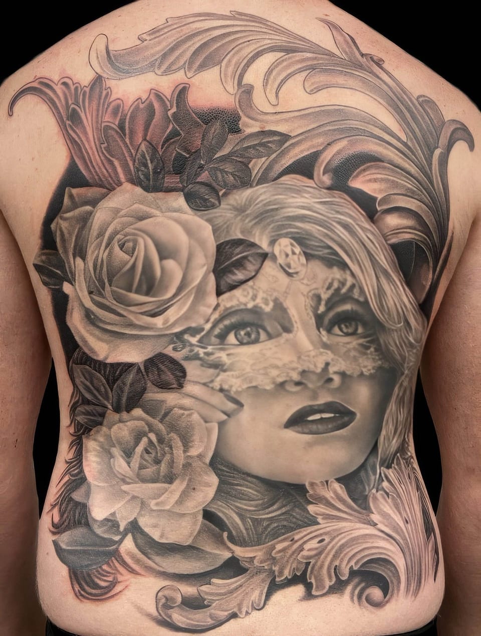 A black and grey realistic woman's face with a lace mask, roses and scrollwork covers a back.