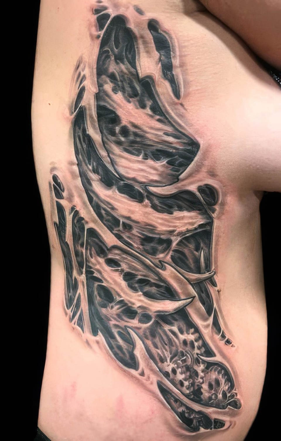 A black and grey bioorganic tattoo with a torn-skin effect covers the side of a torso.