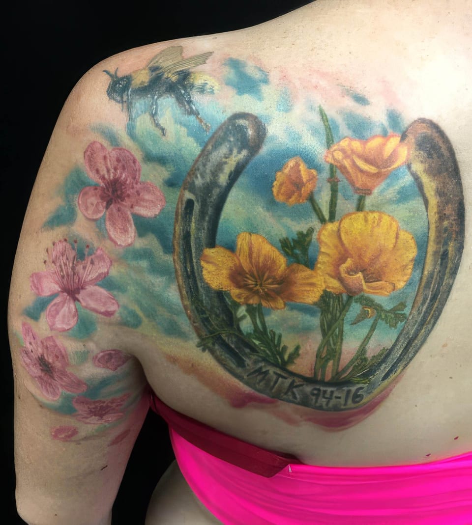 A color realism horseshoe with yellow flowers, pink blossoms and a bee covers the shoulder blade and upper arm.
