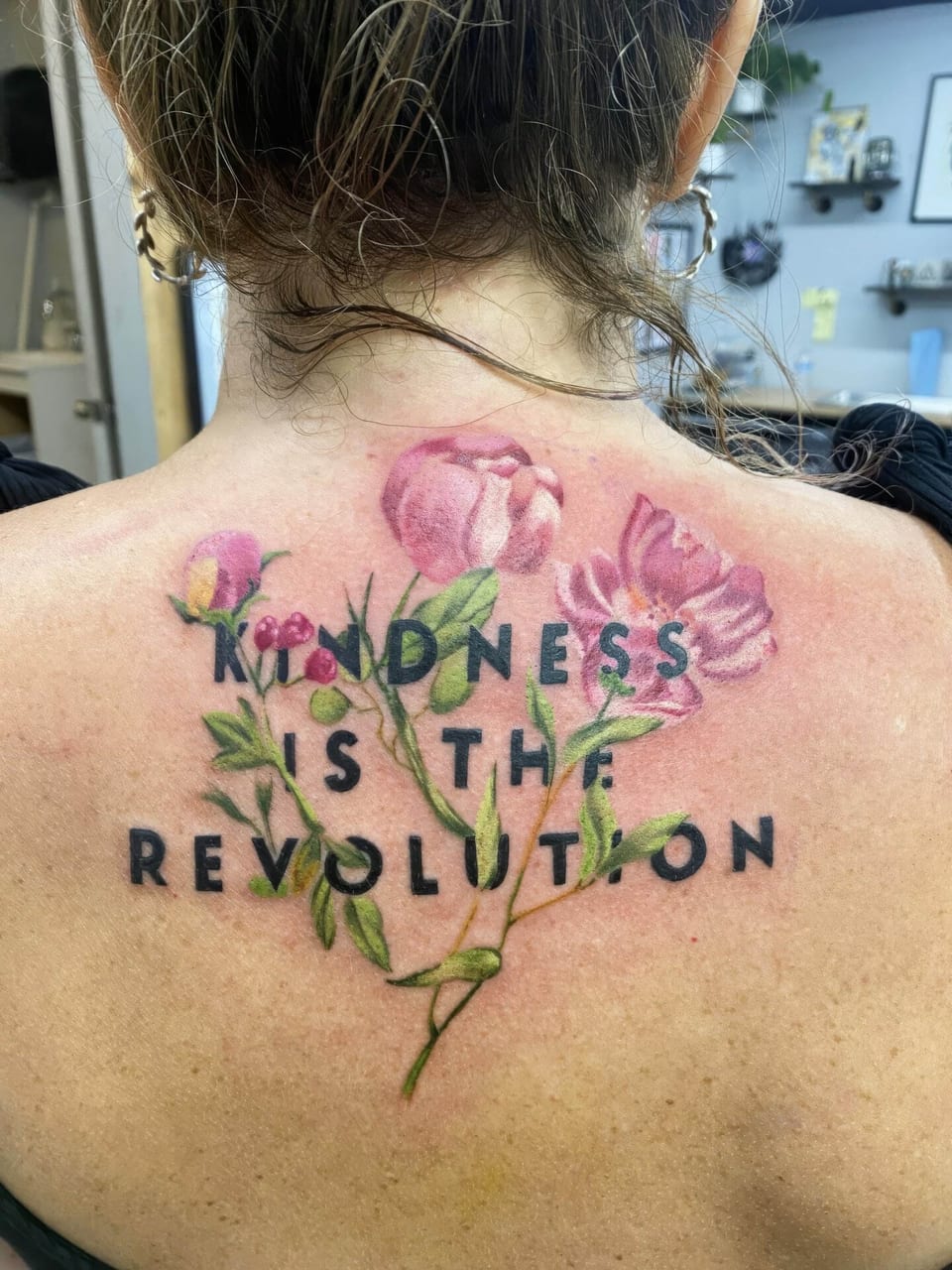 Realistic pink flowers and black lettering reading Kindness is the revolution cover an upper back.