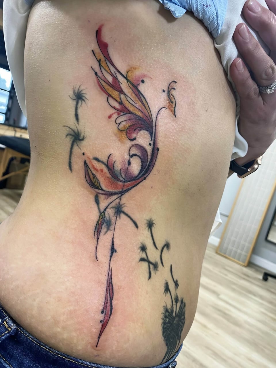 A pink and gold watercolor phoenix with black flourishes covers the ribs beside dandelion seeds.