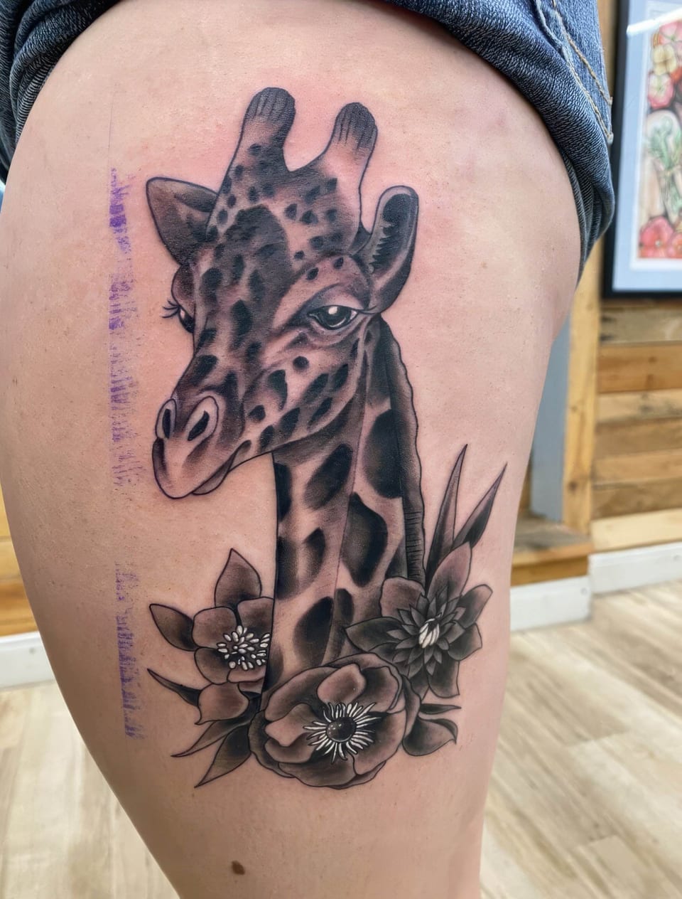 A black and grey neo-traditional giraffe and flowers cover a thigh.