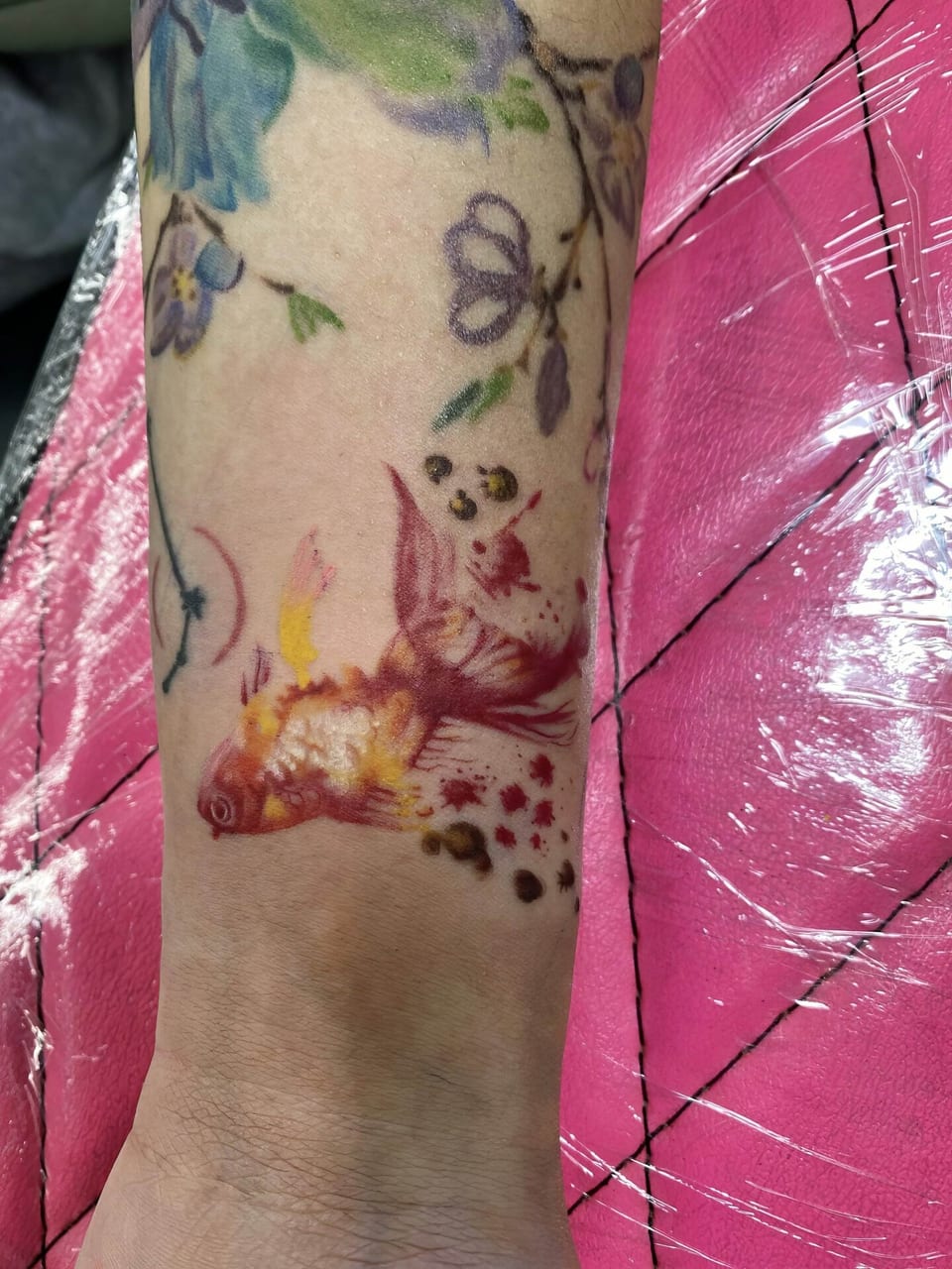 A red and gold watercolor goldfish sits among floral tattoos near a wrist.