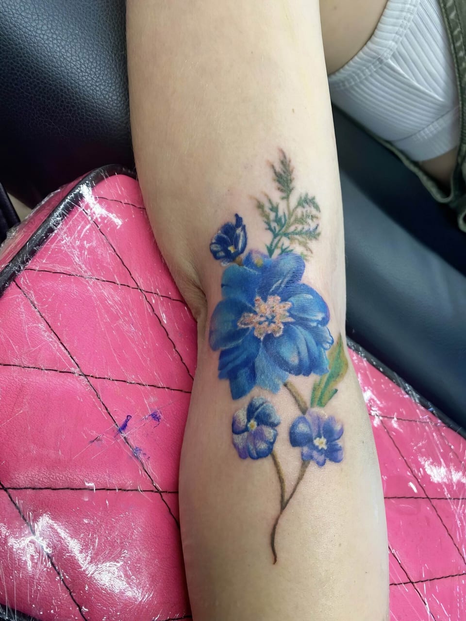 Realistic blue flowers with green leaves and a thin stem cross the inner elbow and forearm.