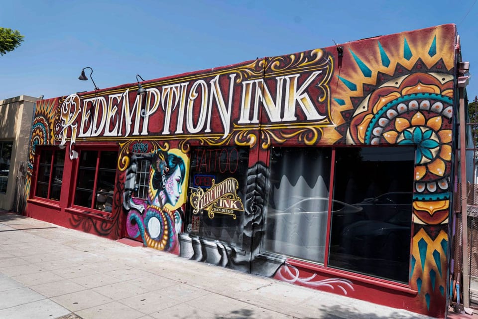 Redemption Ink's red storefront features ornate white lettering, mandala murals and a painted woman beside the door.