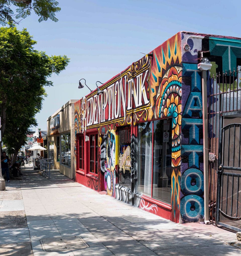 Redemption Ink's colorful storefront and vertical Tattoo lettering face a tree-lined sidewalk.