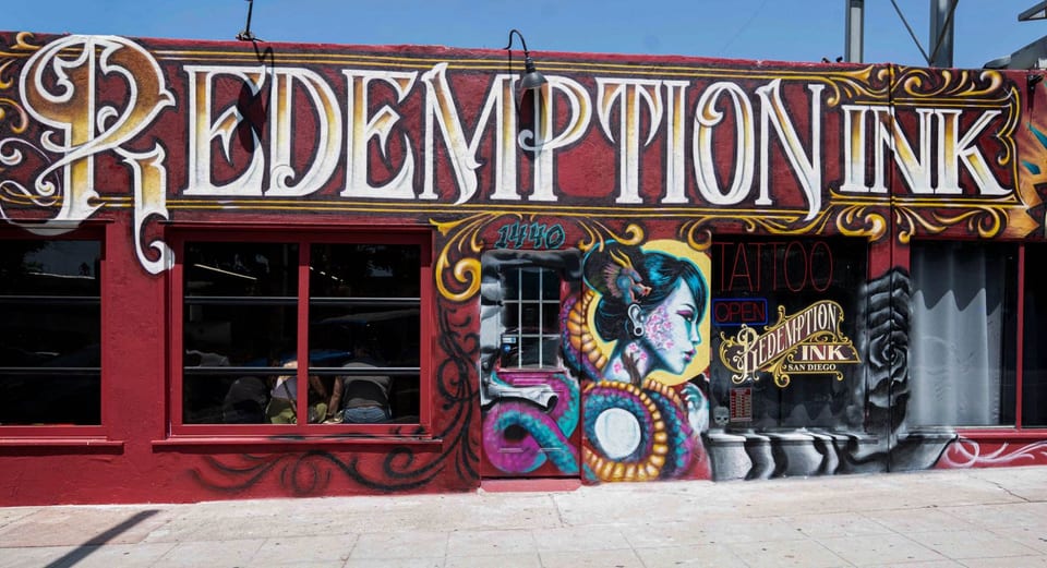 Redemption Ink's storefront has red-framed windows and a colorful woman mural beside the entrance.