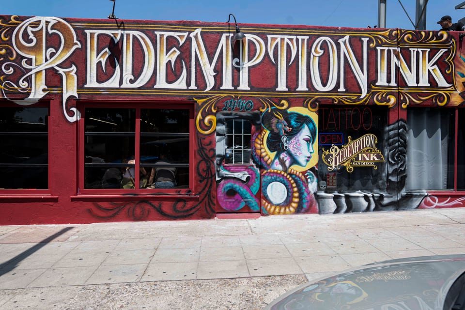 Tighter front view of the Redemption Ink shopfront, with the large painted R, the portrait mural and a car hood below.