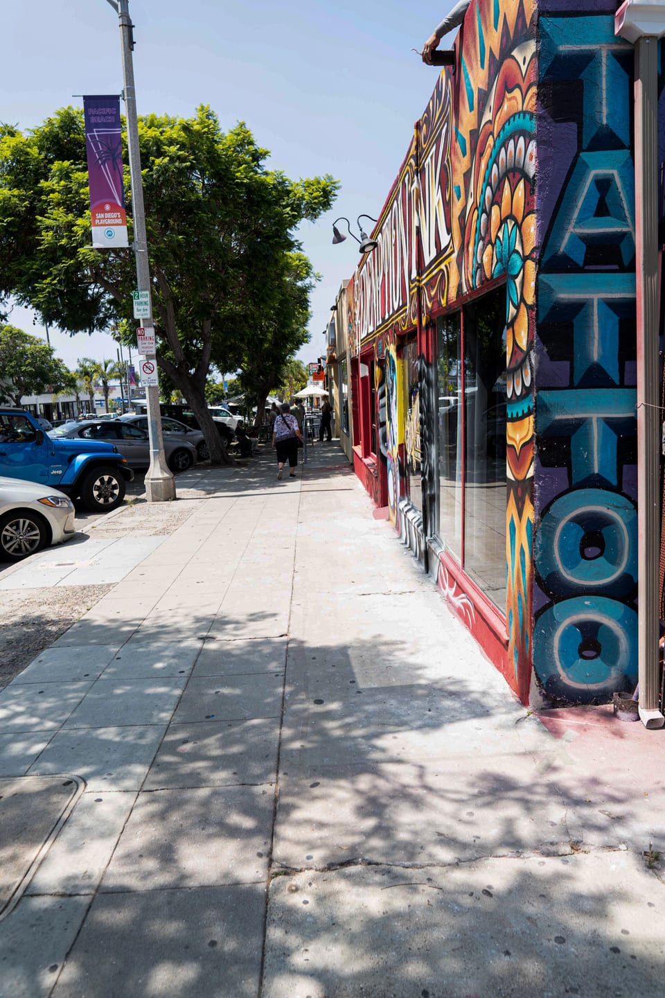 Vertical teal Tattoo lettering faces a tree-lined sidewalk beside Redemption Ink, with a street banner and parked cars.