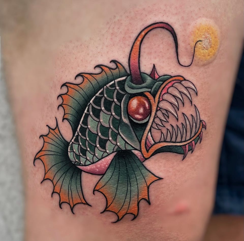 A neo traditional green anglerfish with orange fins and a red eye covers a cropped limb.